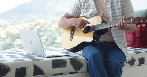 Mature man playing acoustic guitar on window bench with laptop and city view