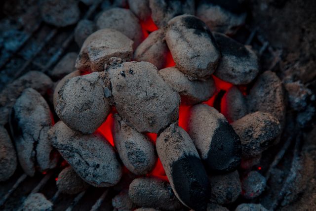 Charcoal briquettes glowing red on a grill, with some covered in ashes, creating a vibrant cooking scene. Ideal for use in materials related to barbecuing, grilling, outdoor cooking, camping experiences, food preparation, and heat-related concepts.