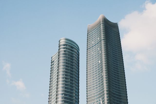 This image captures two modern skyscrapers set against a clear blue sky with some gentle clouds, showcasing contemporary urban architecture. Ideal for use in articles, blogs, or advertisements related to urban development, architecture, real estate, or city profiles.