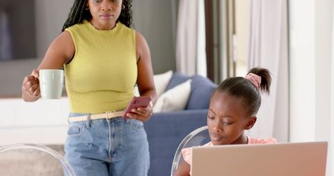 Busy mother multitasking with coffee and phone while daughter doing homework on laptop