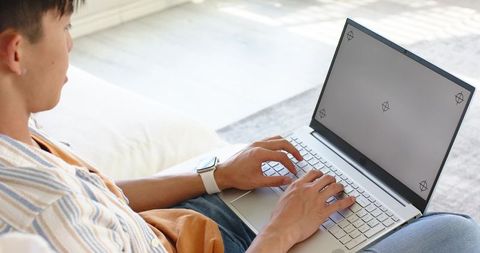 Asian man typing on laptop with smartwatch on sofa in sunlit minimalist office workspace