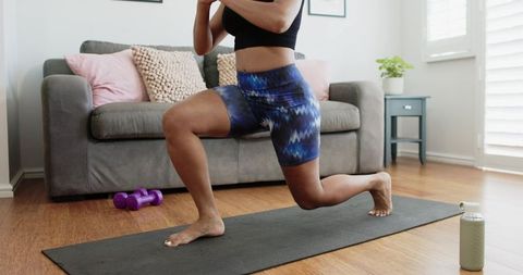 African american woman doing lunges on yoga mat in sunlit living room wearing activewear