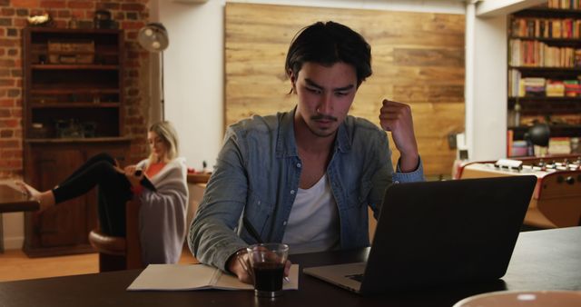 Young man intensely focused on laptop work in a modern, casual home office setting. Open notebook, pen, and glass on desk suggest productivity and focus. Woman in background relaxing on sofa, immersed in her phone. Suitable for illustrating remote work, modern workspace, and multitasking concepts in professional or creative settings.