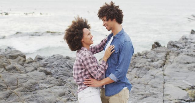 Couple standing on rocky shoreline, sharing a warm embrace and smiling. They are wearing casual clothing, with waves gently crashing in the background. Ideal for promoting romantic getaways, relationship advice blogs, or nature-themed campaigns.