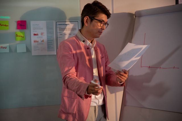 Asian businessman reading a document and drawing a chart on a presentation pad in a modern office. Ideal for use in business presentations, corporate training materials, marketing strategies, and teamwork illustrations.