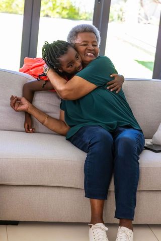 African grandmother hugging granddaughter on sofa in sunlit living room, warm embrace