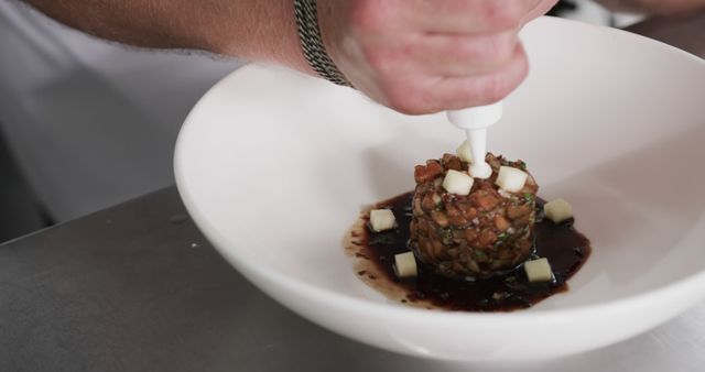 Professional chef meticulously preparing a gourmet tuna tartare dish with fine sauces on a white plate. Ideal for use in content about culinary arts, gourmet cooking, professional kitchens, or fine dining restaurants. Showcases attention to detail and food presentation in the kitchen.