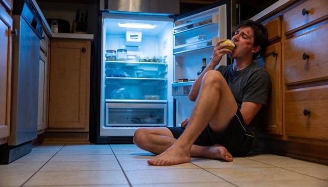 Late-night fridge snack barefoot man eating green apple on kitchen floor by open refrigerator light