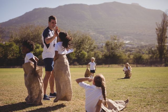 Coach high-fiving schoolgirl during sack race outdoors on a sunny day. Children participating in a fun and competitive activity on a grassy field with mountains in the background. Ideal for use in educational materials, sports and fitness promotions, teamwork and leadership campaigns, and outdoor activity advertisements.