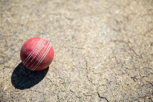 Cricket ball lying on a dry, cracked pitch under bright sunlight. Ideal for use in sports-related content, cricket training materials, or articles about outdoor activities and summer sports.
