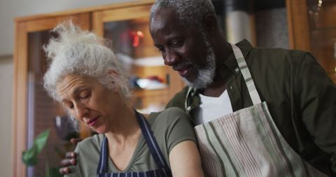 Senior biracial couple bonding in kitchen wearing aprons