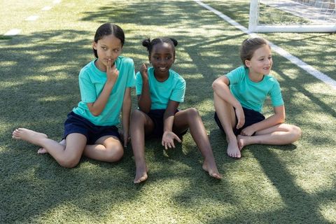 Three young female soccer teammates sitting on turf barefoot wearing teal shirts near goal