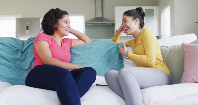Two diverse female friends talking and laughing sitting on couch at home, in slow motion. Free time, friendship and lifestyle concept.