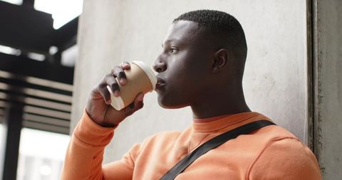 African american commuter sipping coffee by concrete column wearing orange sweater