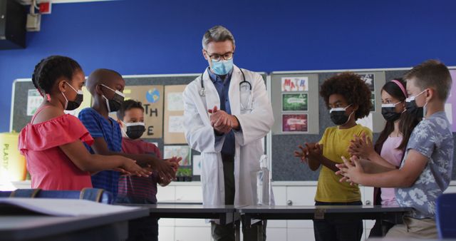 Diverse schoolteacher and schoolchildren standing disinfecting hands, all wearing face masks. children in primary school during coronavirus covid 19 pandemic