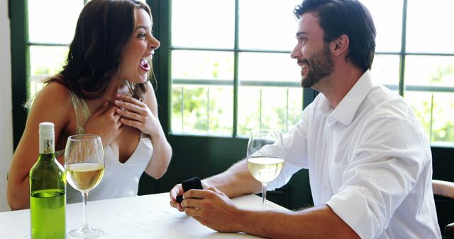 A man is proposing to a Caucasian woman, capturing a moment of surprise and joy during a romantic setting, with copy space. They appear to be young adults enjoying a wine-filled celebration, indicating an engagement or a significant relationship milestone.