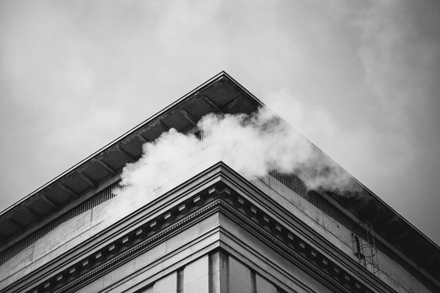 This image features a monochrome view of a building's corner, with steam rising against a cloudy sky. Architectural elements and geometric lines are emphasized by the black and white contrast. The steam adds a dynamic element to the still composition. Ideal for use in urban and architectural projects, as well as for themes involving industrial scenery or modern cityscapes.