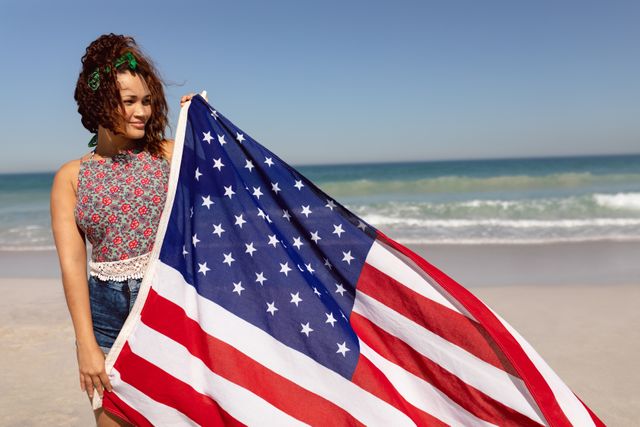 Biracial woman with curly brown hair and light brown skin holding American flag at beach, looking thoughtful. She is wearing a floral top and denim shorts. Ideal for themes of patriotism, summer activities, national pride, and American holidays such as Independence Day or Memorial Day.