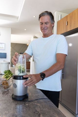 Mature man preparing green smoothie with personal blender in modern home kitchen