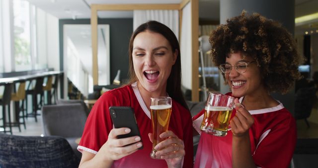 Two women are smiling and holding beer glasses while looking at a smartphone in a bar. The setting suggests a casual and friendly environment where people can enjoy a good time and socialize. Great for use in content related to social gatherings, nightlife, friendship, leisure activities, and modern lifestyle.