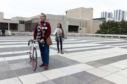 Mature couple walking urban checkerboard plaza with road bike and coffee by brutalist architecture