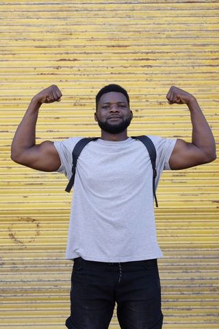African man flexing biceps with backpack against yellow shutter urban fitness confidence