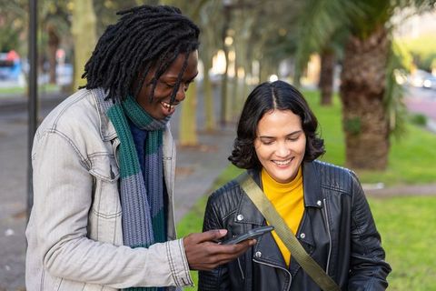 Smiling diverse friends checking smartphone on tree-lined park path, urban casual lifestyle