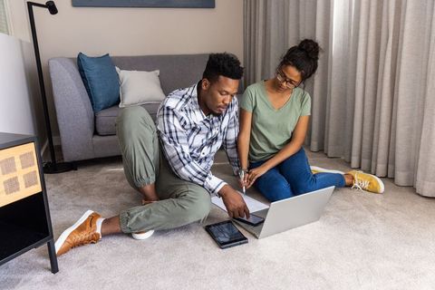 Diverse couple reviewing finances on laptop and tablet while sitting on living room floor