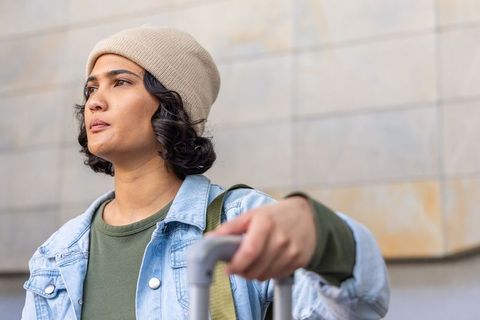Young woman standing with suitcase wearing denim jacket and beanie, looking ahead in city