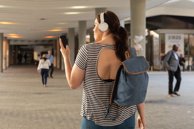 Rear view of curvy Caucasian woman out and about in the city streets during the day, using her smartphone wearing headphones and carrying a backpack with modern architecture in the background