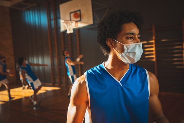 Basketball player wearing face mask practicing with team in sunlit indoor court. Ideal for use in articles about sports during COVID-19, health and safety in athletics, or team training sessions. Can be used in promotional materials for sports facilities, health guidelines, or pandemic-related sports activities.