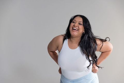 Curvy woman laughing and leaning forward in white ribbed tank, neutral gray background