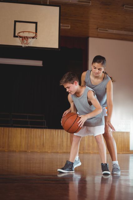 Two school kids are playing basketball in a gymnasium. One child is dribbling the ball while the other is closely guarding. This image is ideal for illustrating youth sports, physical education programs, teamwork, and active lifestyles in educational settings.