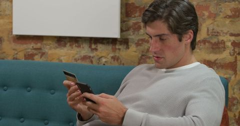 Young Man Shopping Online with Smartphone and Credit Card on Sofa