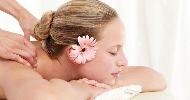 A young Caucasian woman enjoys a relaxing massage in a serene spa environment, with copy space. Gentle hands work to relieve tension in her shoulders, enhancing the tranquil experience with a touch of floral beauty.