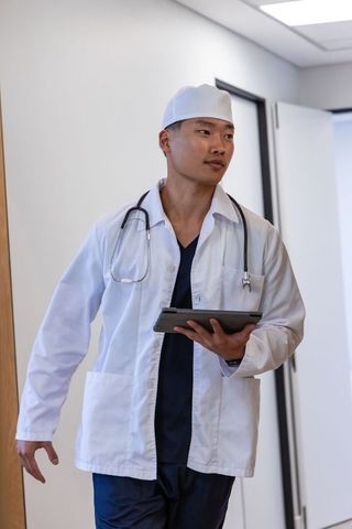 Asian male physician walking clinic corridor holding tablet and stethoscope