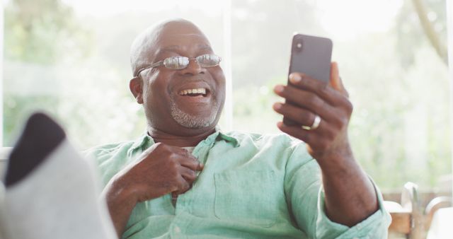 Happy african american senior man sitting with feet up making image call with smartphone, laughing. retirement lifestyle, spending time alone at home.