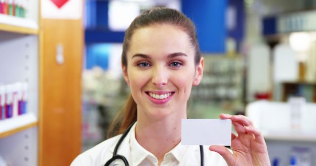 Young female doctor smiling and holding a blank business card in a medical office. Ideal for use in advertisements, healthcare campaigns, medical website designs, and professional branding materials.