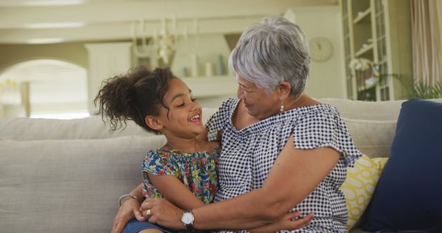 Happy biracial granddaughter and grandmother sitting on couch embracing at home. Childhood, family, home, togetherness, domestic life and senior lifestyle, unaltered.