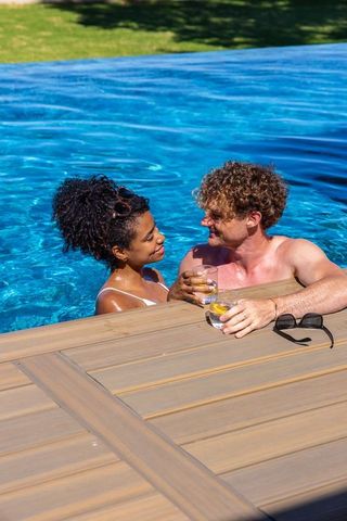 Interracial couple relaxing in backyard pool leaning on wooden deck holding lemon drinks