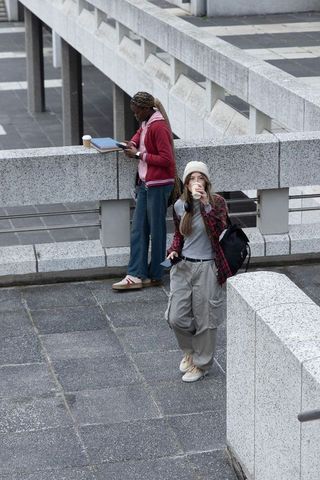 Diverse young women standing sipping coffee and using phone and tablet at urban plaza