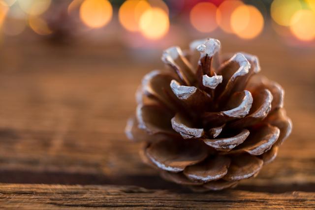 This image features a pine cone with fake snow on a wooden plank, set against a background of bokeh lights. Ideal for use in holiday-themed marketing materials, Christmas cards, festive blog posts, and seasonal social media content. The rustic and natural elements make it perfect for promoting eco-friendly holiday decorations.