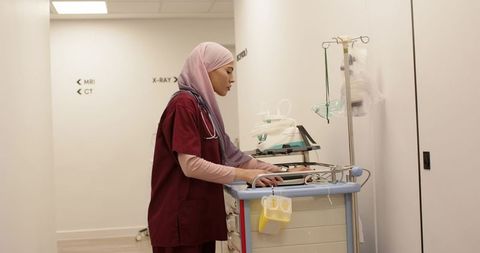 Hijab-wearing nurse adjusting crash cart monitor in radiology corridor, clinical readiness