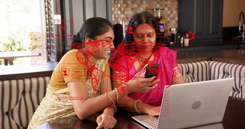 Two indian women in sarees using smartphone and laptop at kitchen table with data overlay