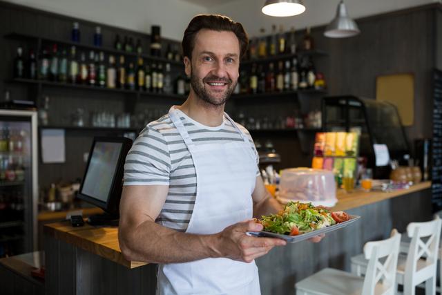 Waiter holding tray of fresh salad in restaurant, smiling at camera. Ideal for use in hospitality industry promotions, restaurant marketing materials, healthy eating campaigns, and customer service training resources.