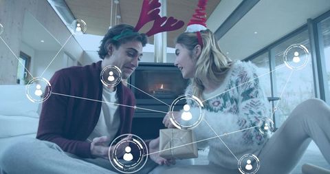 Two women exchanging holiday gift while wearing reindeer antlers by cozy fireplace