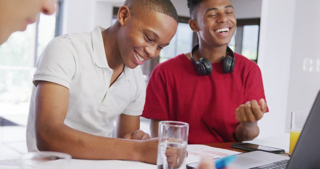 Two teenage boys, one wearing a white shirt and the other a red shirt with headphones, are sitting at a table engaged in studying and laughing together. Glasses of water and a laptop are on the table, indicating a casual and comfortable environment. This image is suitable for educational materials, online articles about teenage life and study habits, and promotional content for academic resources.