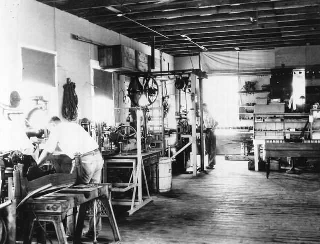 This historic scene captures the interior of Dr. Robert Goddard's rocket lab at Mescalero Ranch, New Mexico during the early 1930s. It shows two workers, A. Kisk and C. Mansur, among an array of machinery and tools used in pioneering rocketry experiments. This image is perfect for illustrating the history of space exploration, innovation, and the early days of NASA's rocketry advancements.