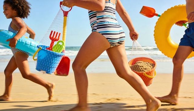 Kids running along sandy shoreline carrying beach buckets, inflatables and shovels