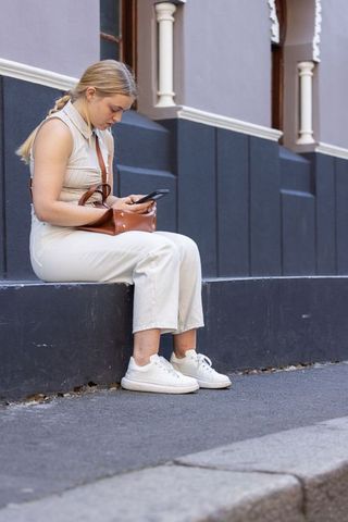Young woman sitting on city ledge checking smartphone, casual outfit, white sneakers